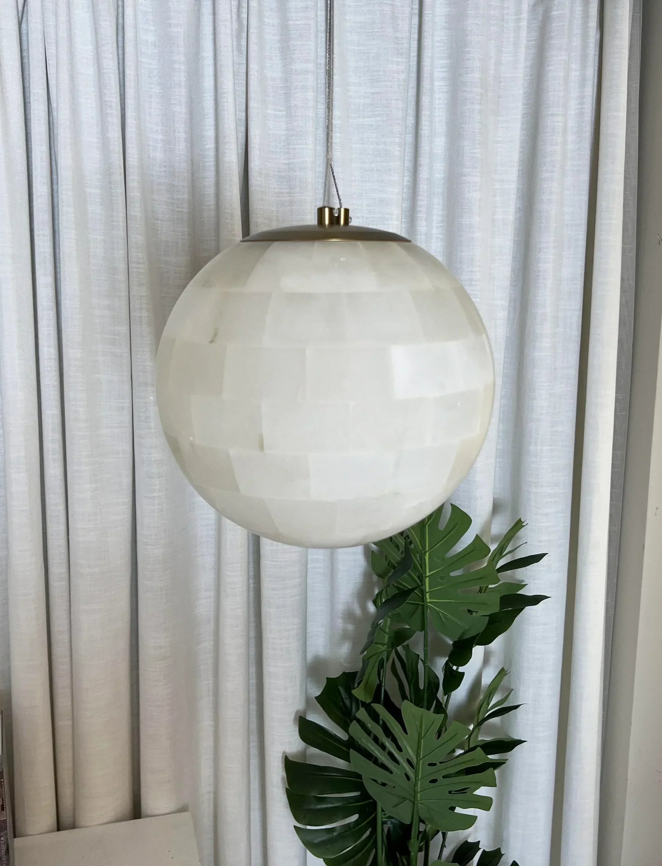 Modern alabaster ball pendant light with brass accents hanging in front of white curtains and green leaves