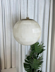 Modern alabaster ball pendant light with brass accents hanging in front of white curtains and green leaves