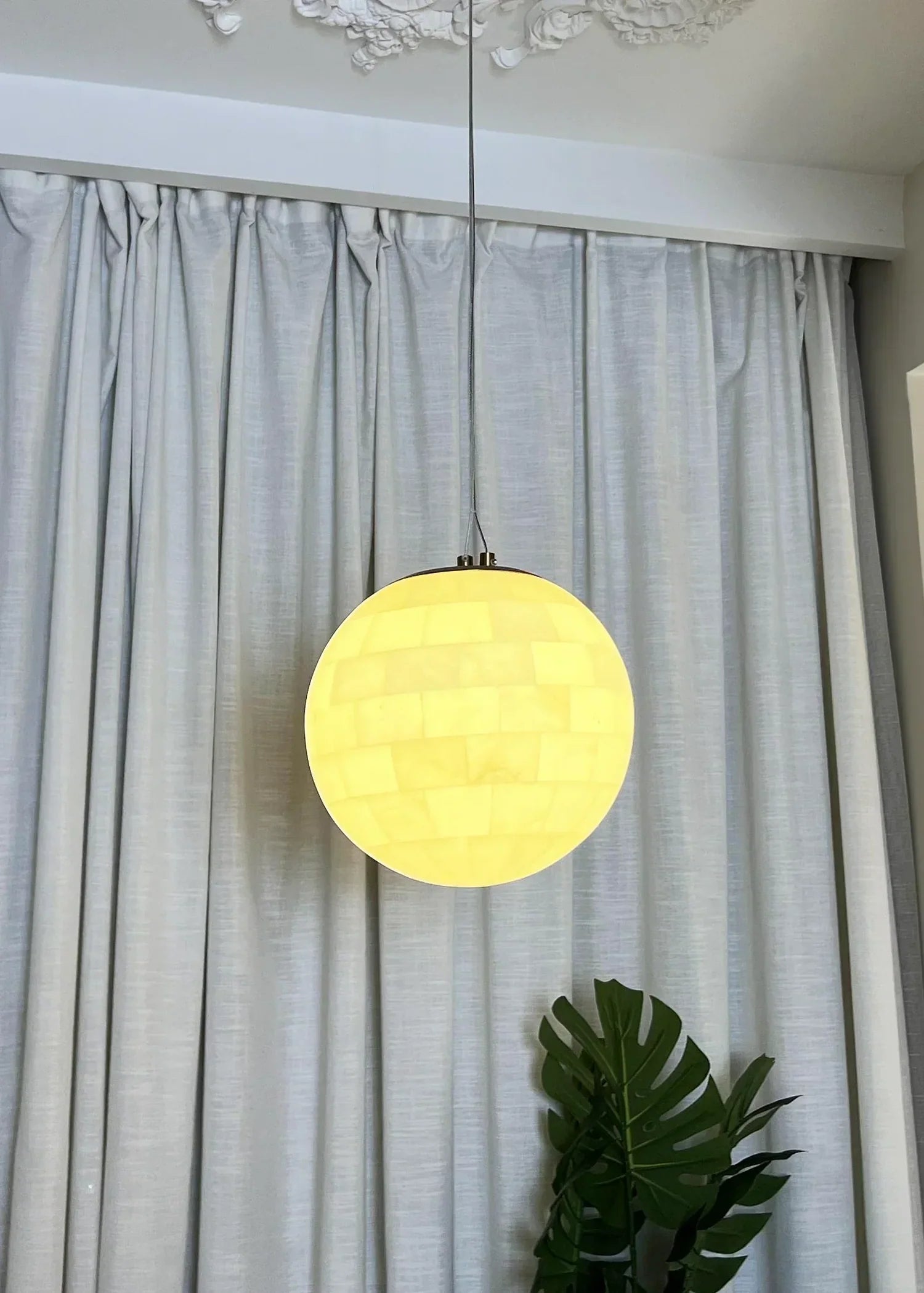 Alabaster ball pendant light glowing warmly, hanging in front of white curtains with green plant below