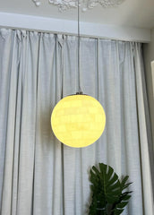 Alabaster ball pendant light glowing warmly, hanging in front of white curtains with green plant below
