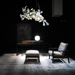 Modern living room setup with white ginkgo leaf chandelier, dark wood table with glowing lantern, cushioned chair and ottoman on a gray rug