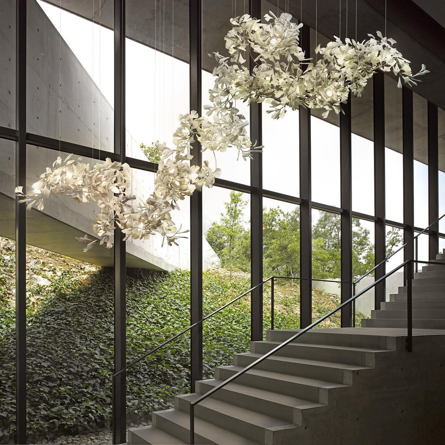 Modern white ginkgo leaf chandelier with gold accents hanging above staircase in building with large windows showing outdoor greenery