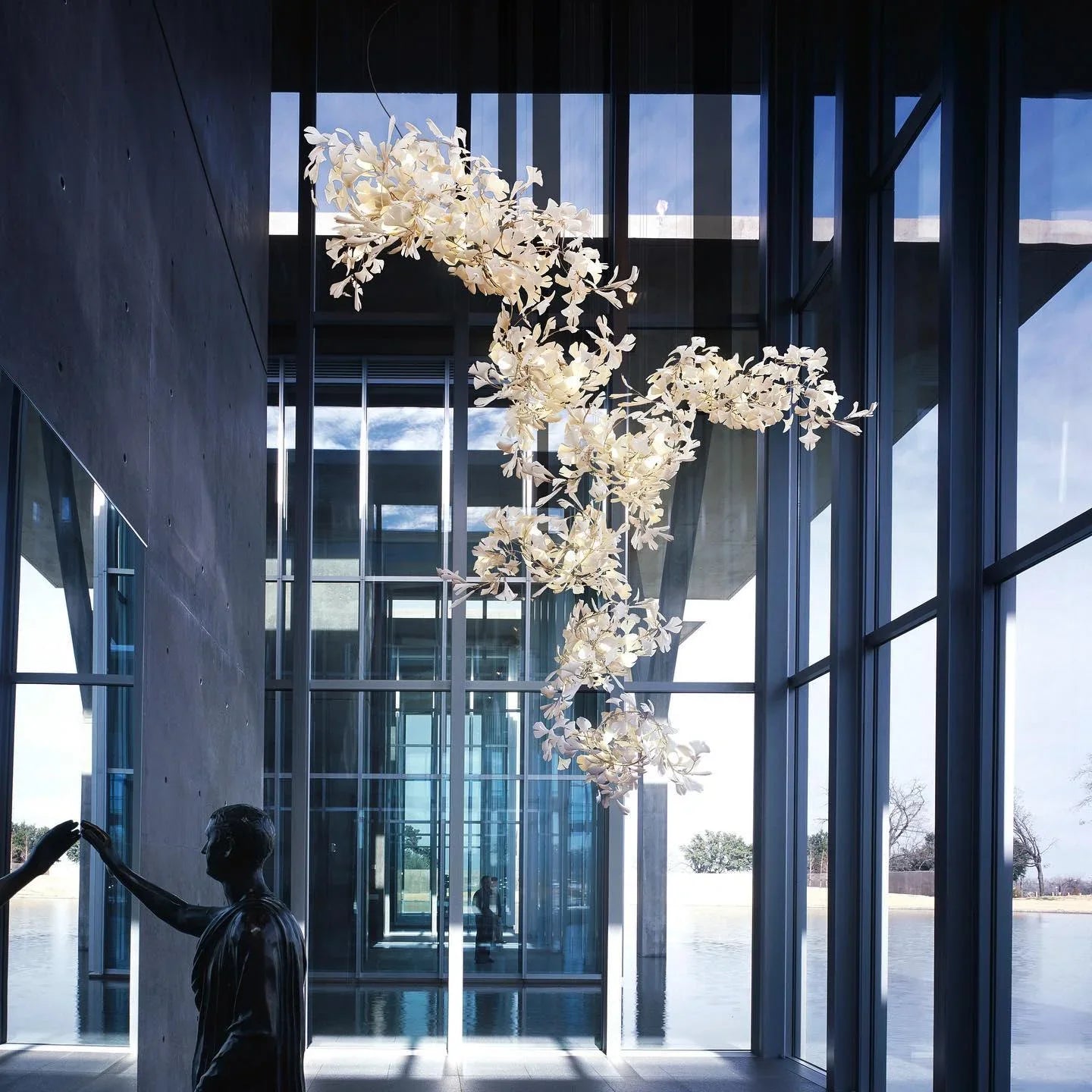 Modern gold and white gingko leaf chandelier in high-ceilinged glass room with reflective sculpture