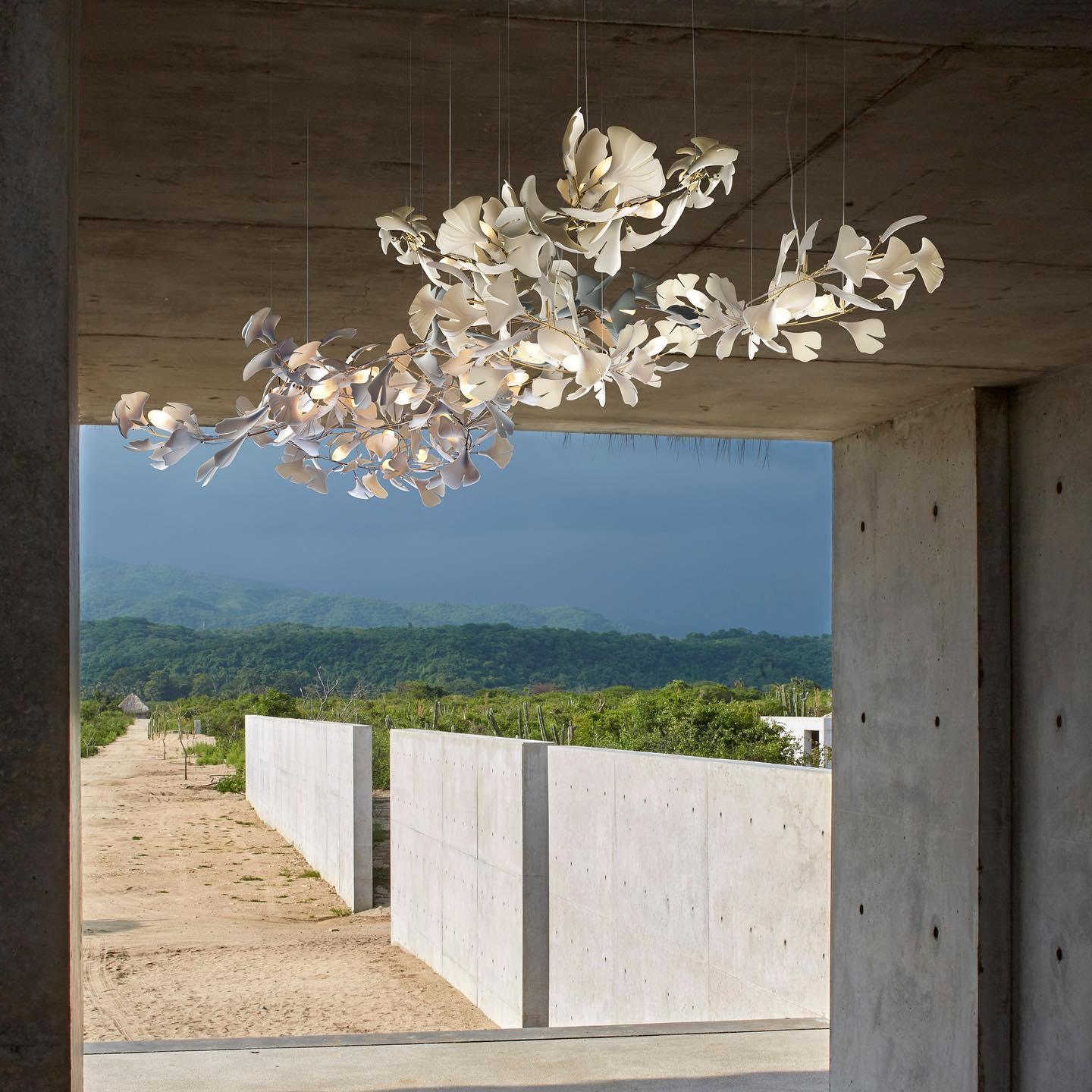 Modern gold and white gingko leaf chandelier suspended in a concrete room with outdoor nature view