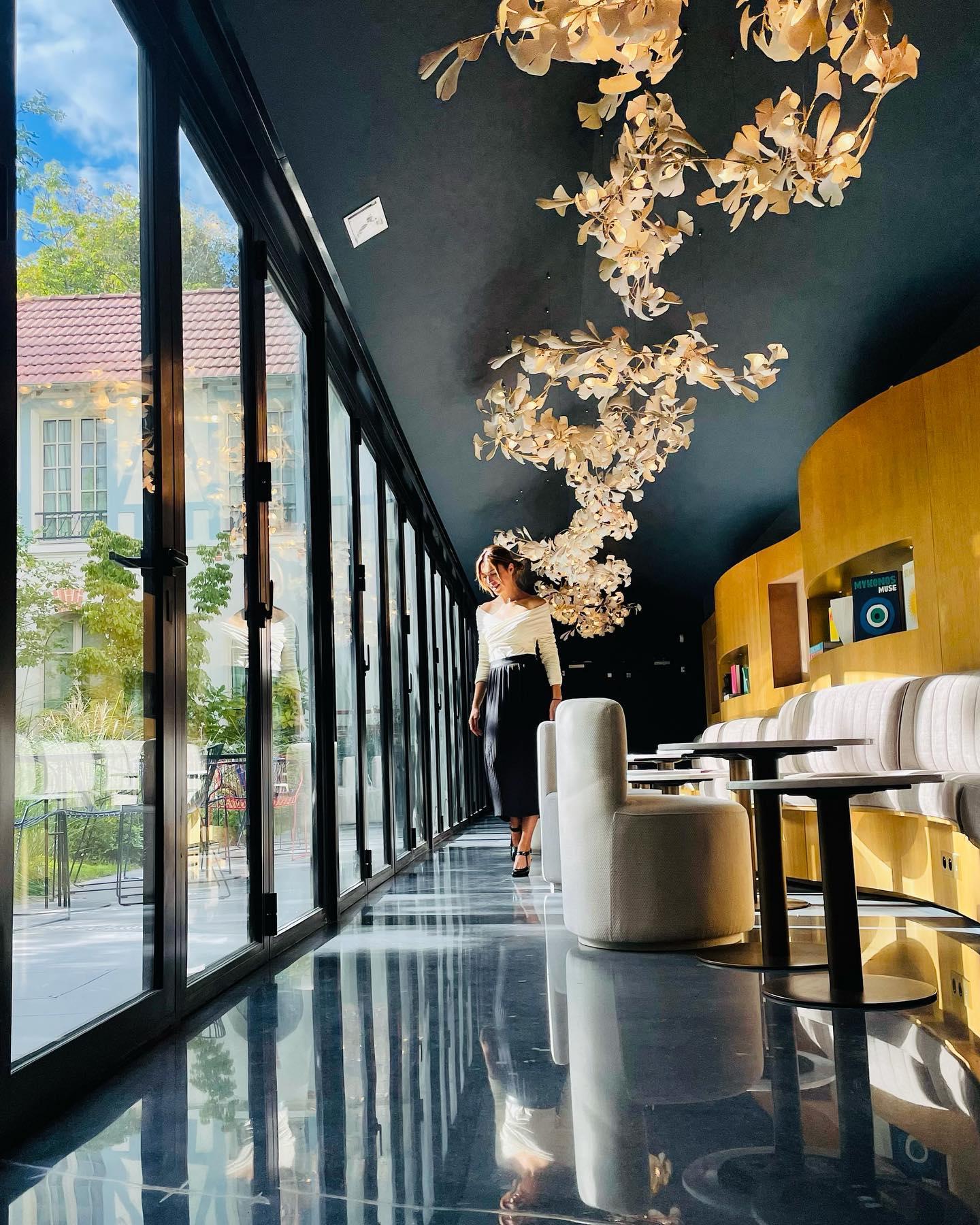 Modern indoor hallway with floral gold-white gingko chandeliers, black ceiling, glass doors, and a woman walking
