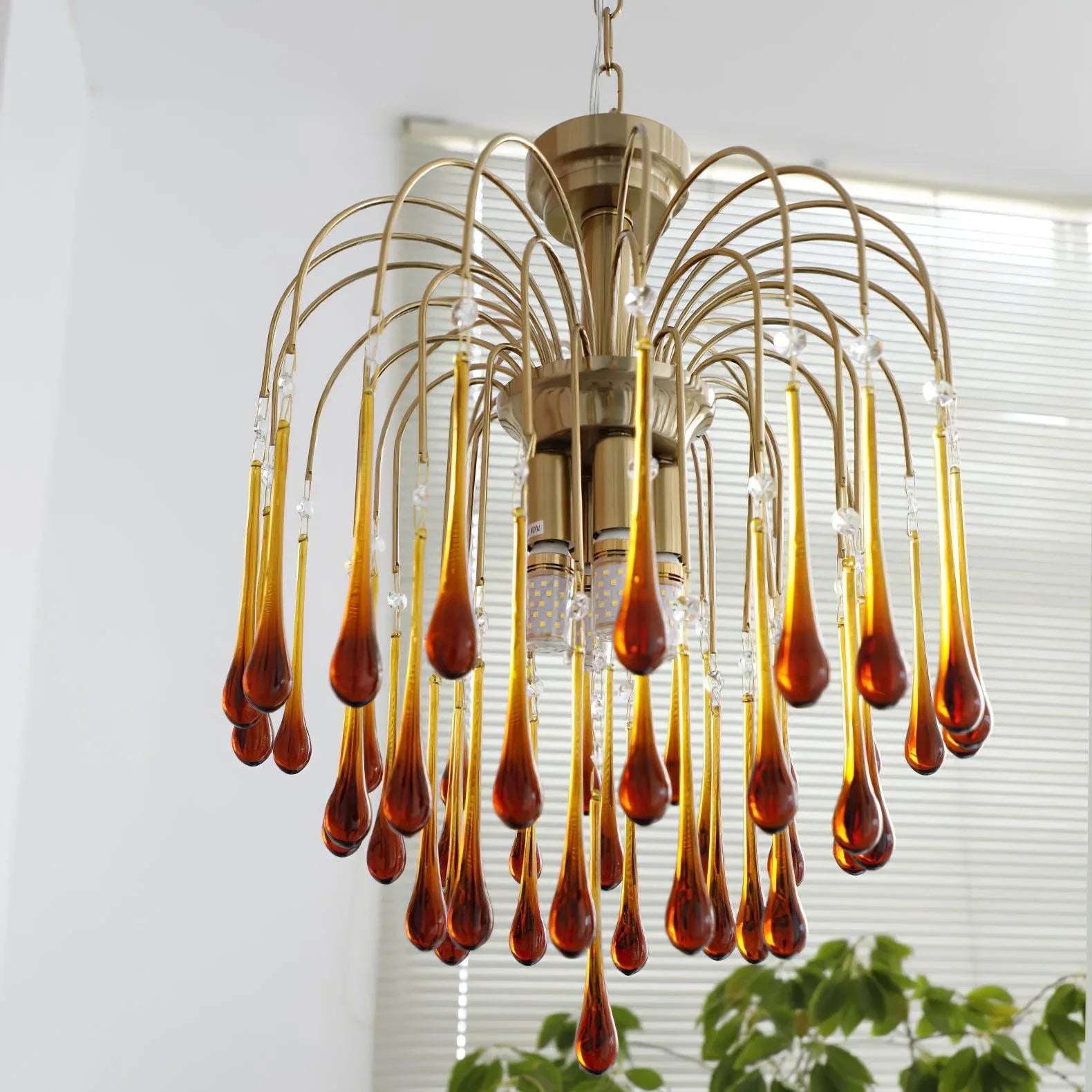 Modern brass chandelier with amber glass teardrop pendants hanging in a bright room with window blinds and plants