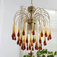 Modern brass chandelier with amber glass teardrop pendants hanging in a bright room with window blinds and plants
