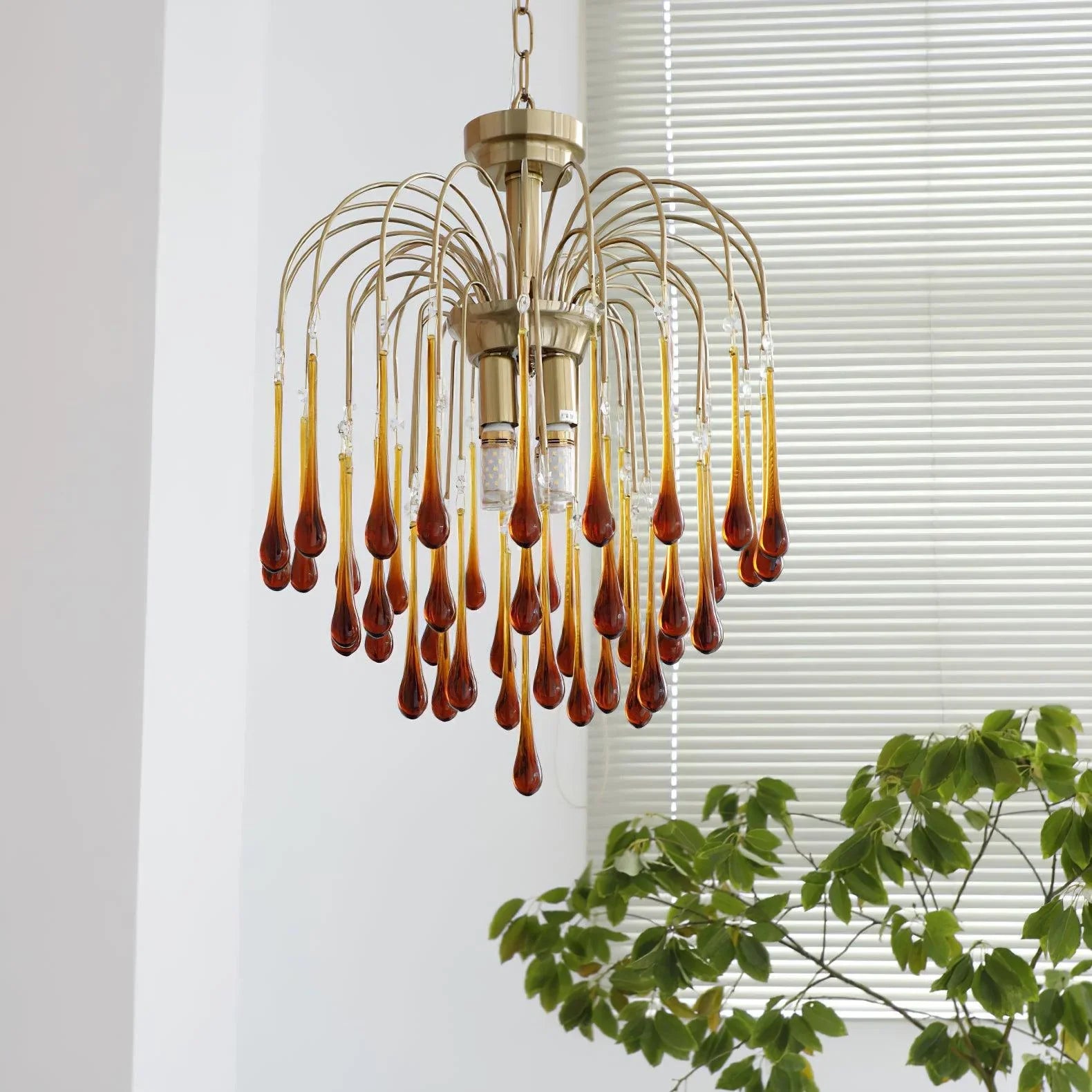 Modern brass chandelier with amber glass teardrop pendants hanging in a bright room with window blinds and green plant
