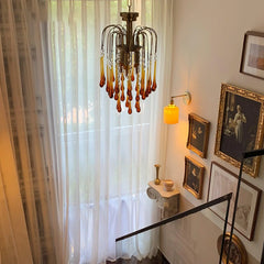Modern brass chandelier with amber teardrop crystals hangs in elegant stairwell with sheer curtains and vintage framed art