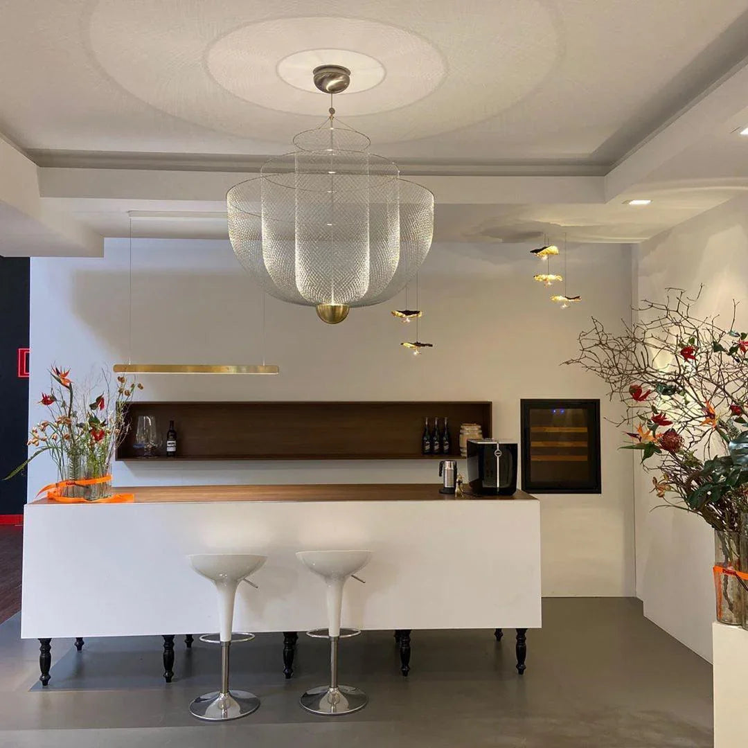 Modern kitchen bar area with white counter, two white bar stools, mesh chrome pendant lamp, and decorative plants