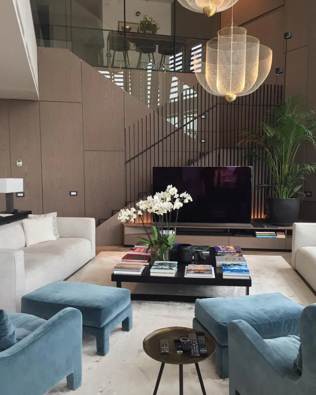 Modern living room with large chandelier, beige sofas, blue velvet chairs, black coffee table with books, and large plant