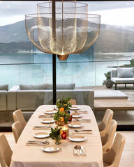 Modern dining room with white tablecloth, set table, and large gold mesh pendant chandelier overlooking lake view
