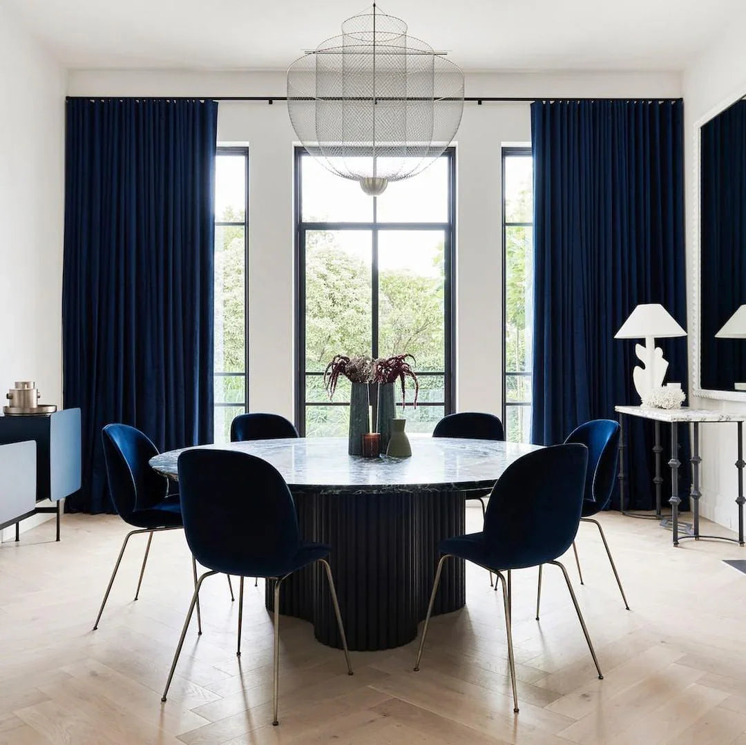 Modern dining room with round marble table, dark blue velvet chairs, large windows, blue curtains, and stylish mesh pendant lamp