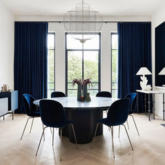 Modern dining room with round marble table, dark blue velvet chairs, large windows, blue curtains, and stylish mesh pendant lamp