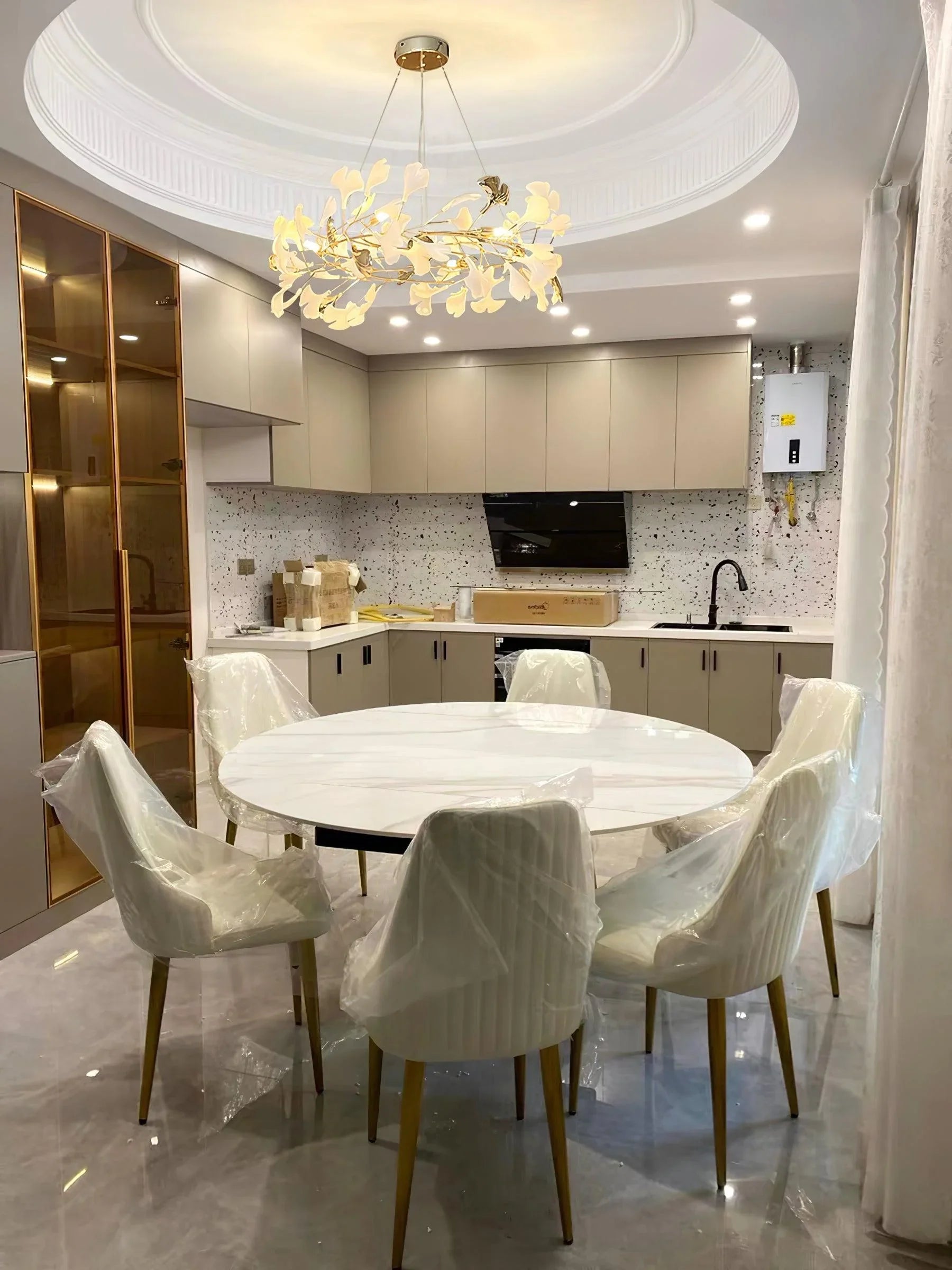 Modern dining area with round white marble table, six white chairs with wooden legs, gold and white gingko leaf chandelier, and beige kitchen cabinetry