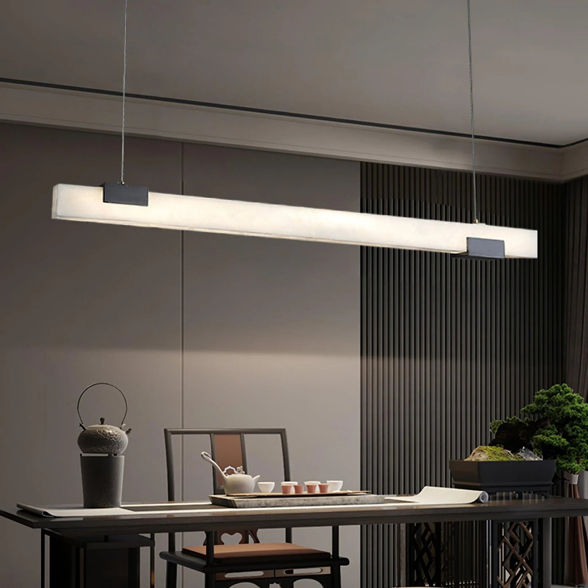 Modern linear alabaster pendant lamp with warm light hanging above a dining table with tea set and decorative plants