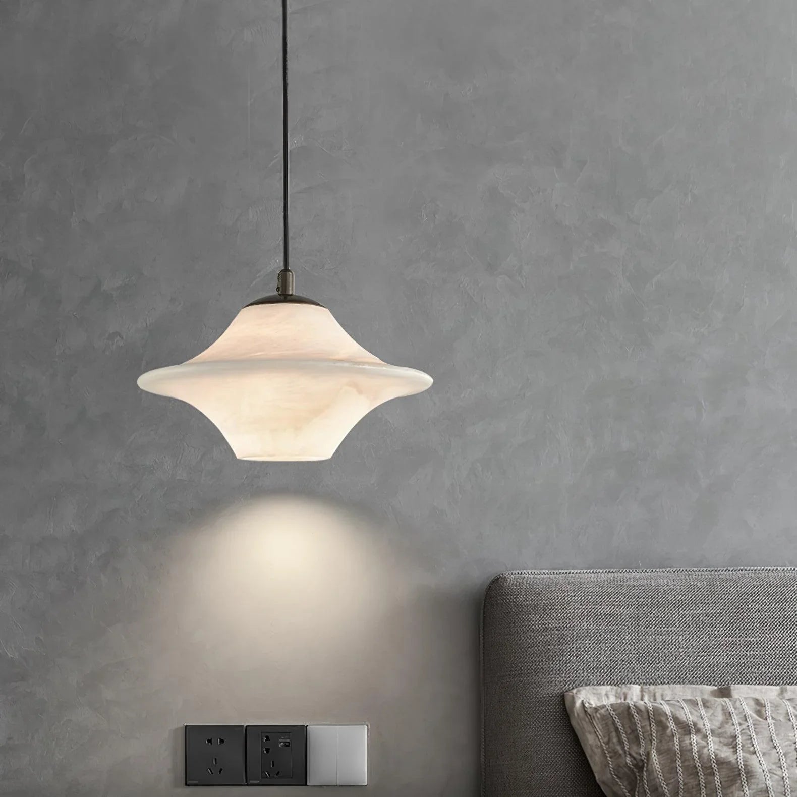 Modern black and white saucer alabaster pendant lamp hanging over a gray textured wall and bed headboard