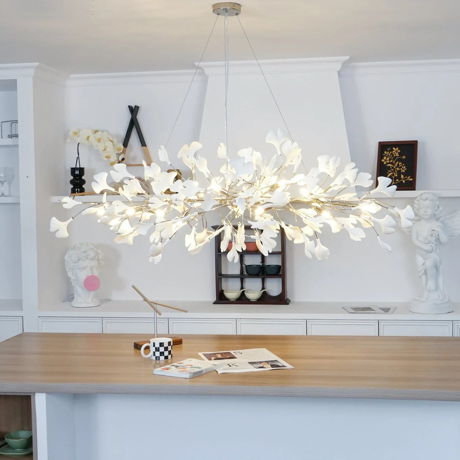 Modern gold and white ginkgo leaf chandelier pendant lamp hanging over wooden dining table in stylish room with decorative sculptures