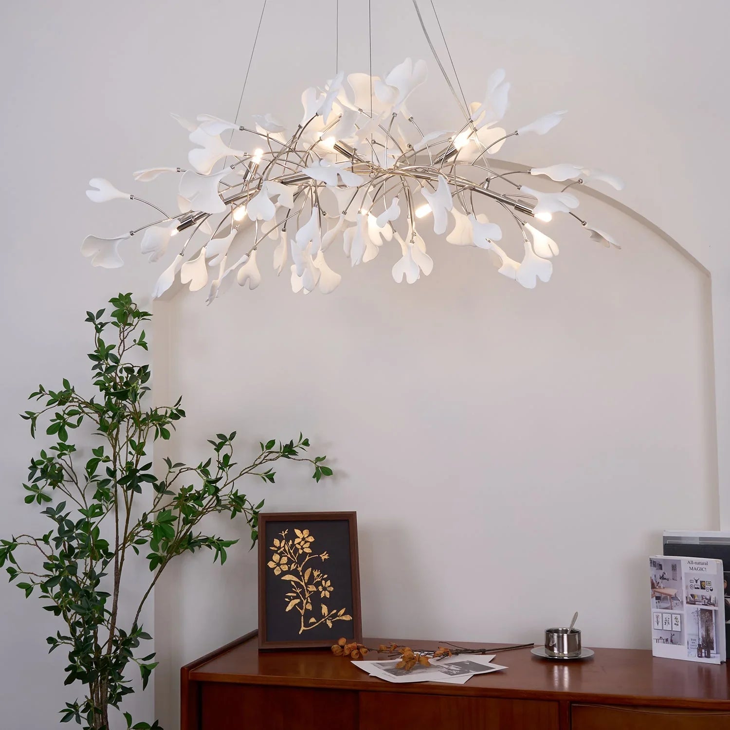 Modern white ginkgo leaf chandelier pendant lamp hanging over wooden sideboard with green plant and framed art