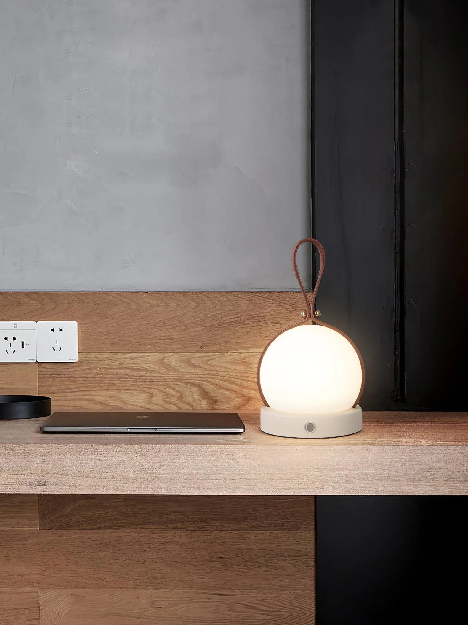 Modern spherical table lamp with brown handle on wooden desk, next to closed laptop, YH Lamps