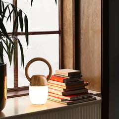 Modern wooden table lamp with circular handle and warm light by window, next to stacked books