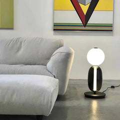 Modern table lamp with glowing round white shade and dark base on floor next to light gray sofa and colorful wall art