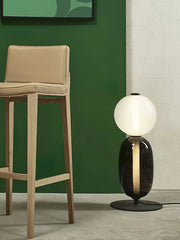 Modern table lamp with candied glass base and white globe shade beside wooden stool against green wall
