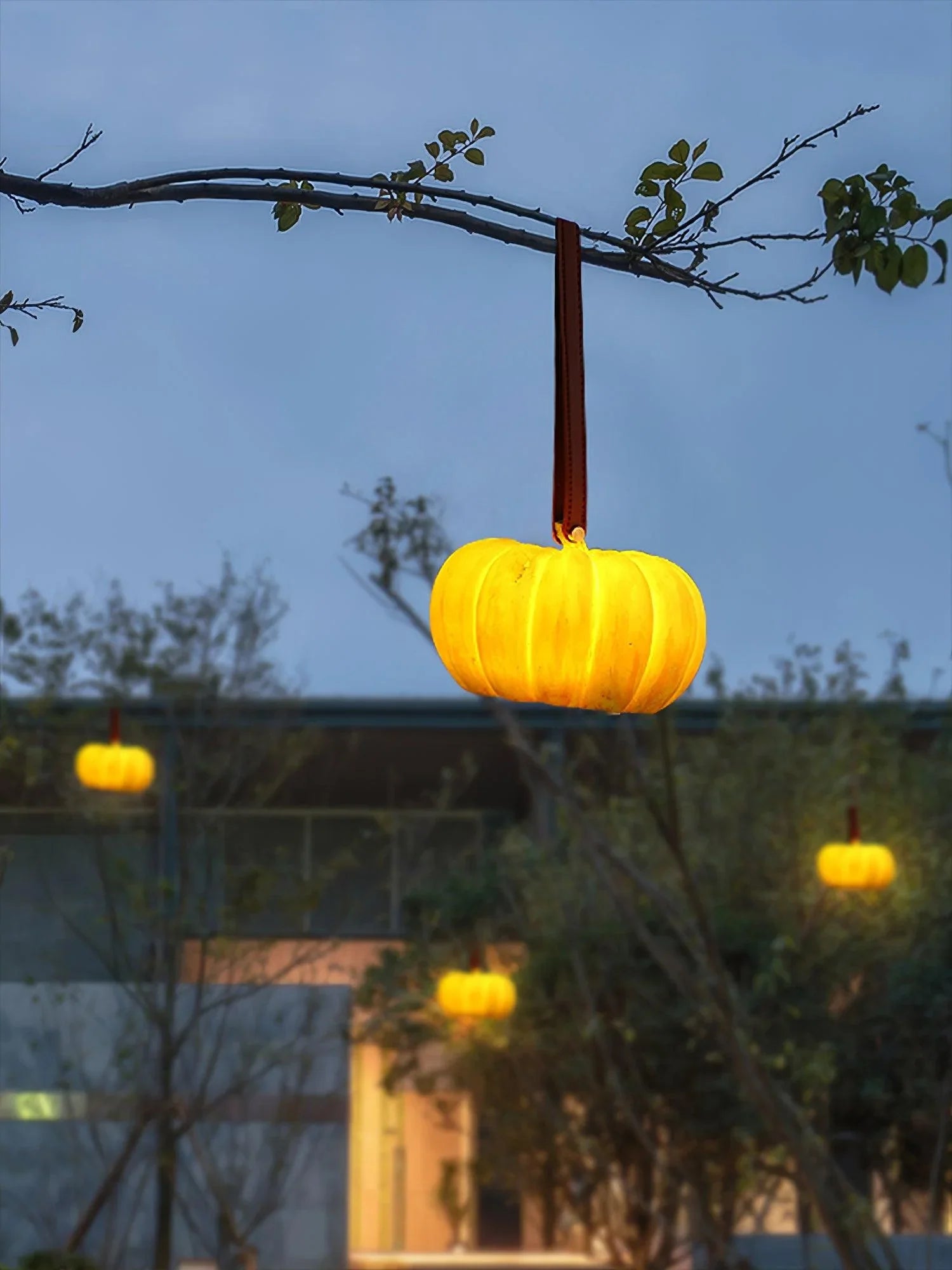 Yellow pumpkin-shaped portable table lamp hanging from a tree branch outdoors at dusk