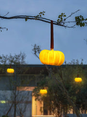 Yellow pumpkin-shaped portable table lamp hanging from a tree branch outdoors at dusk