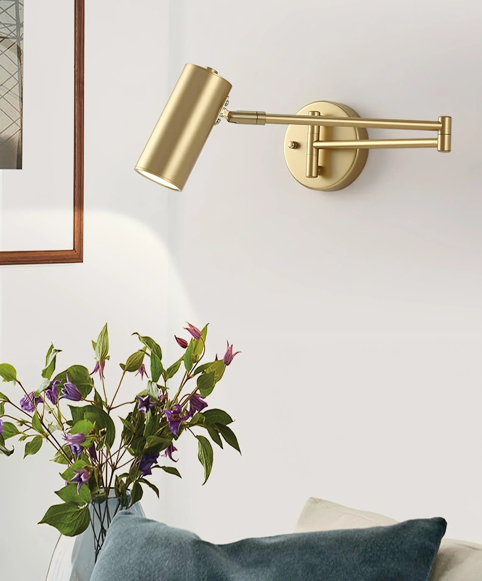 Gold swing arm wall lamp with warm light mounted on white wall above couch and next to vase with purple flowers