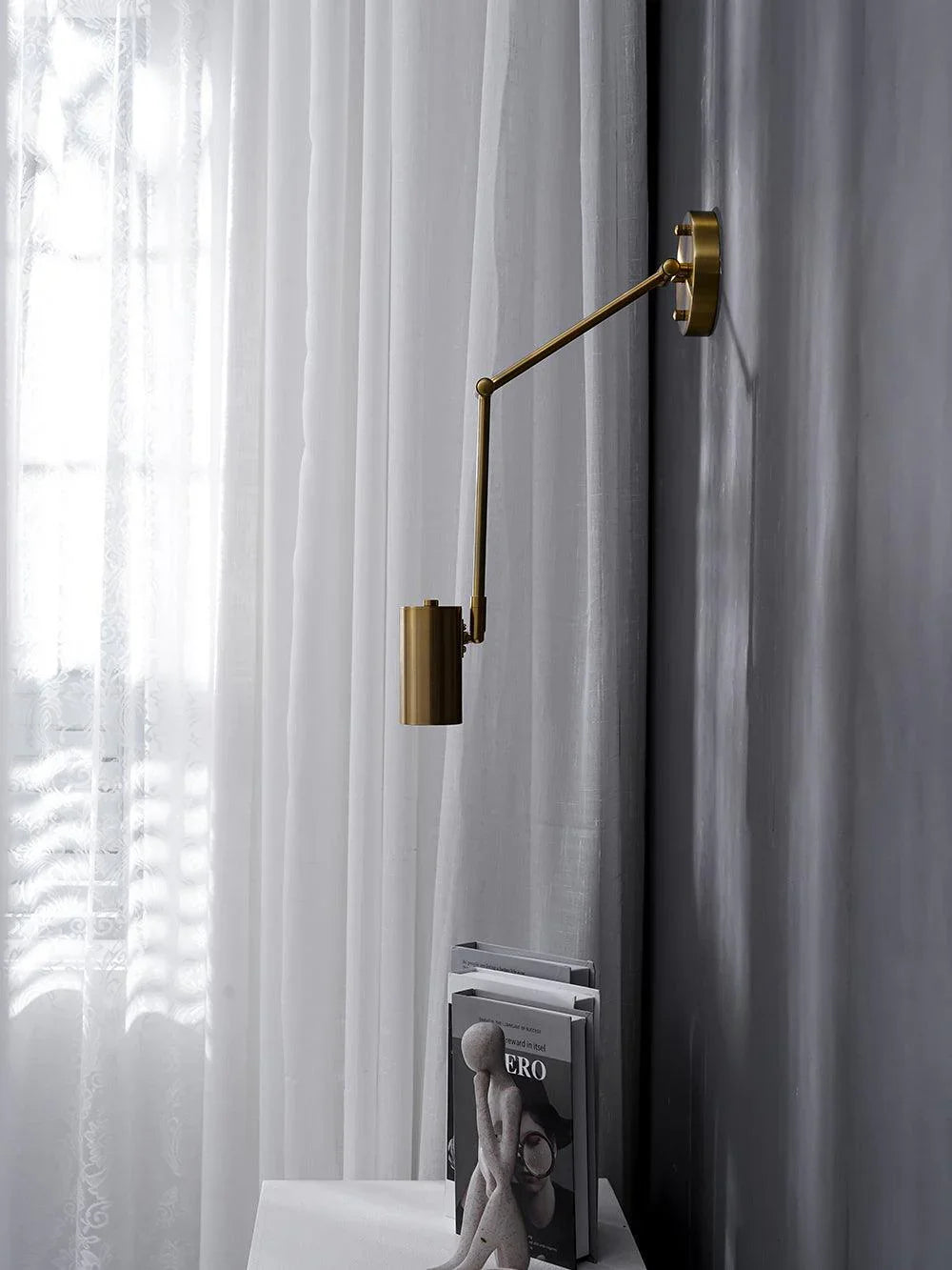 Modern brass swing arm wall lamp with wall switch illuminating gray wall near sheer white curtains and books on table