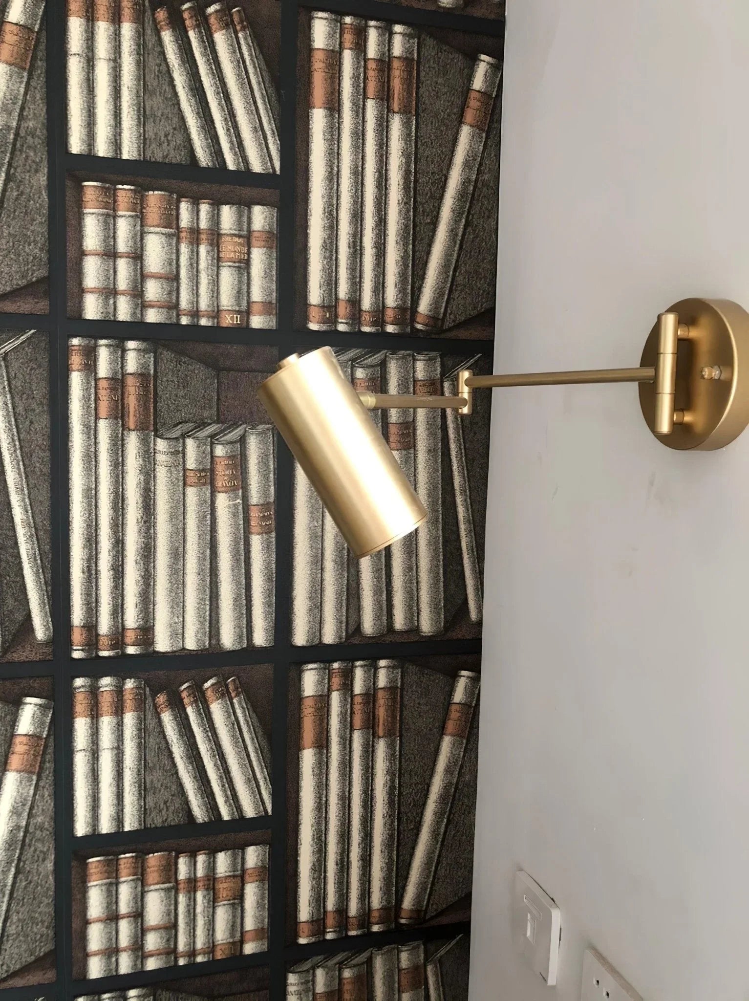 Modern gold swing arm wall lamp mounted next to bookshelf wallpaper with vintage book design