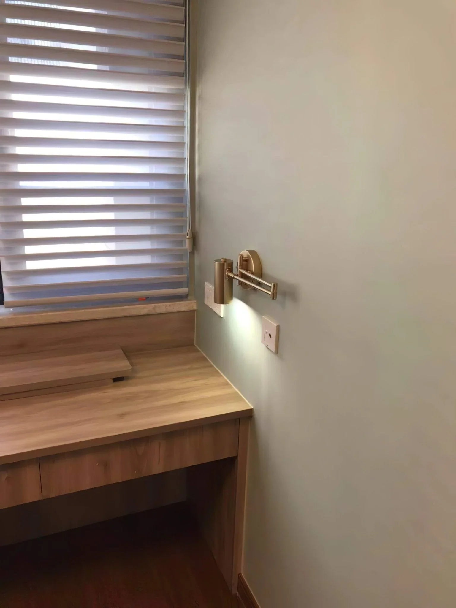 Modern gold swing arm wall lamp with warm light above wooden desk near window blinds