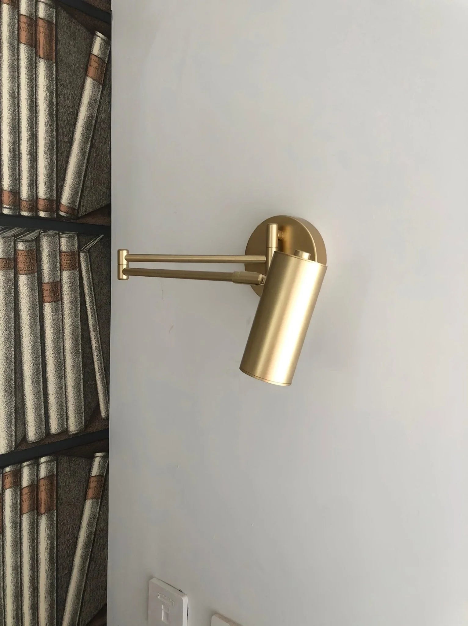 Gold swing arm wall lamp with cylindrical shade mounted on a white wall next to bookshelf wallpaper