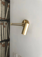 Gold swing arm wall lamp with cylindrical shade mounted on a white wall next to bookshelf wallpaper
