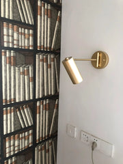 Modern gold swing arm wall sconce mounted on white wall next to bookshelf wallpaper and electrical outlets