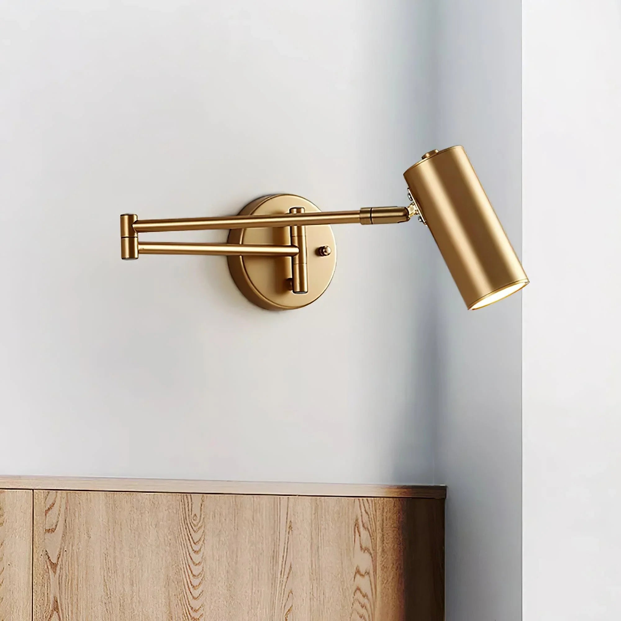 Gold swing arm wall sconce with warm light mounted on white wall above wooden surface